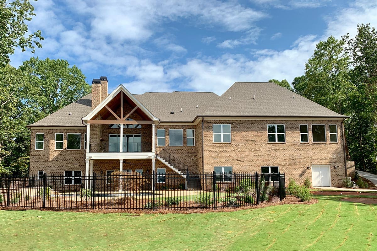 House plan exterior, two-story brick home with exposed rafter trusses, covered balcony, and black metal fence.