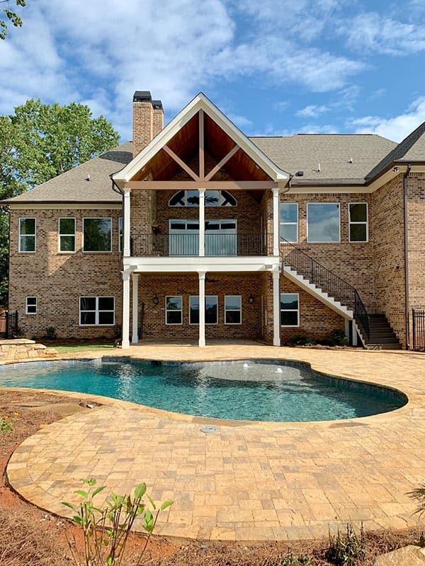 House plan exterior: Two-story brick home with covered upper deck, exposed rafter tails, and a kidney-shaped pool.
