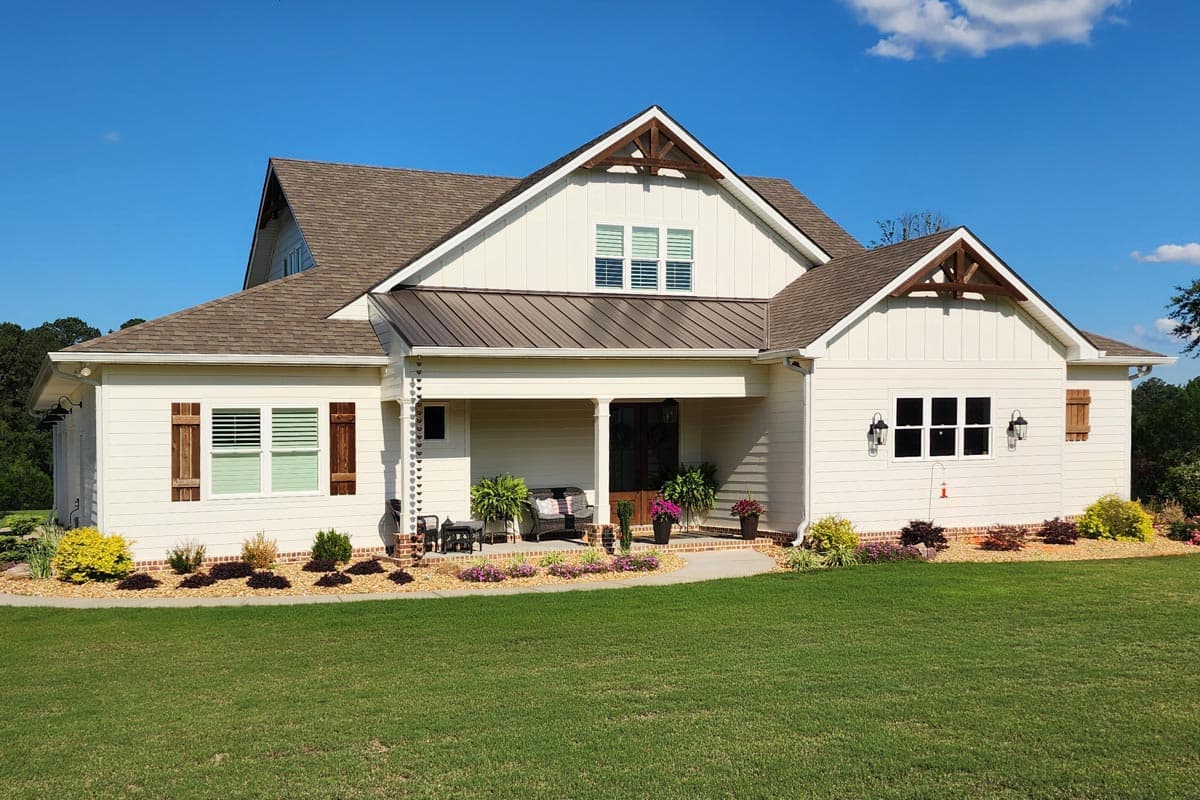 A large white farmhouse with a brown roof and a covered front porch. The home has a manicured lawn and landscaping under a blue sky.
