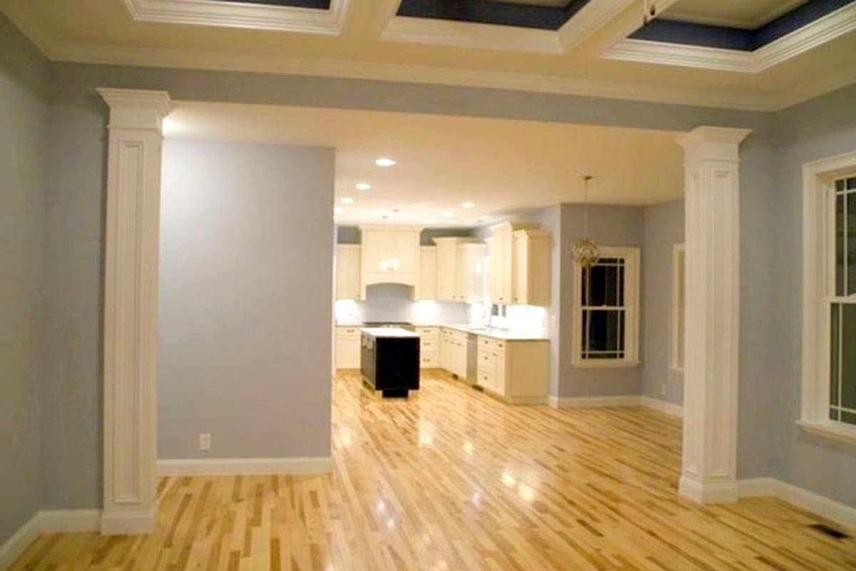 Interior view of a kitchen with cream cabinets, a dark island, and hardwood floors, opening to a living area with columns.