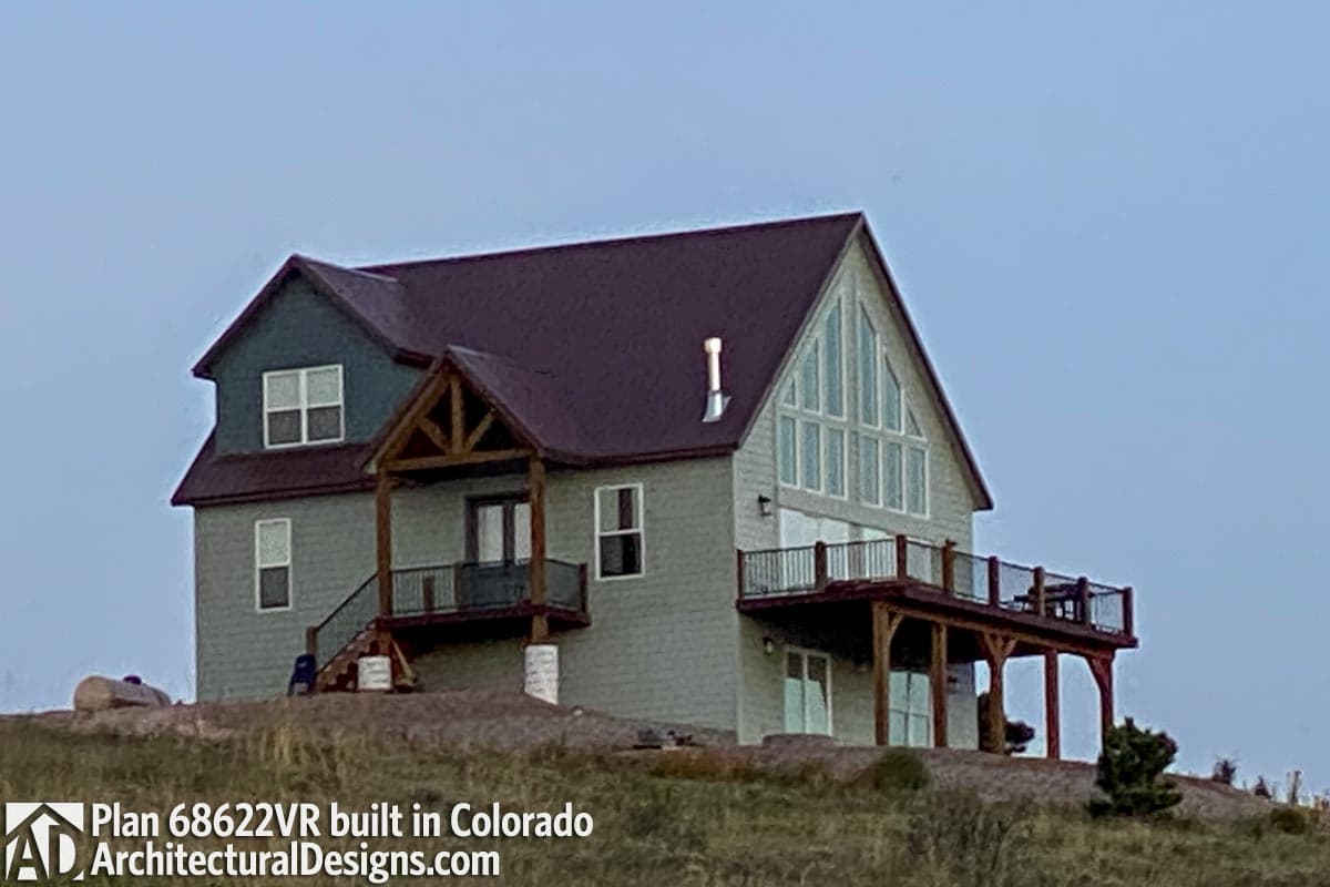 House plan exterior with A-frame gable, large windows, two-story construction, prominent deck, and covered porch.