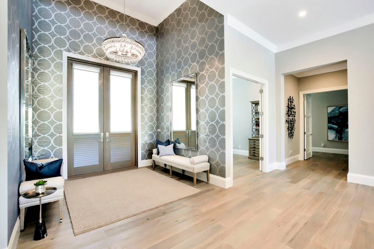 Foyer with double doors, patterned wallpaper, chandelier, bench, and wood floors.
