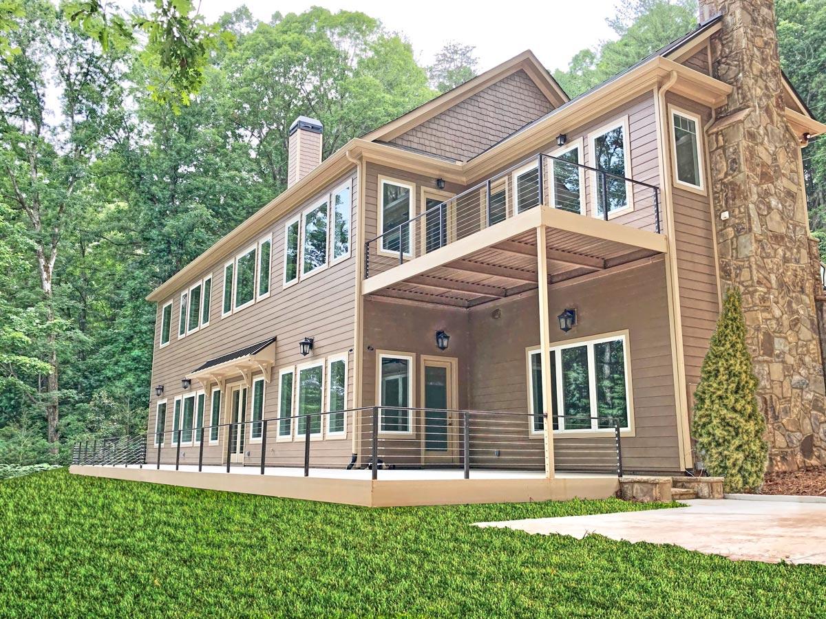 House plan exterior: Tan siding, multiple stories, large stone chimney, upper balcony, and covered lower patio with railing.
