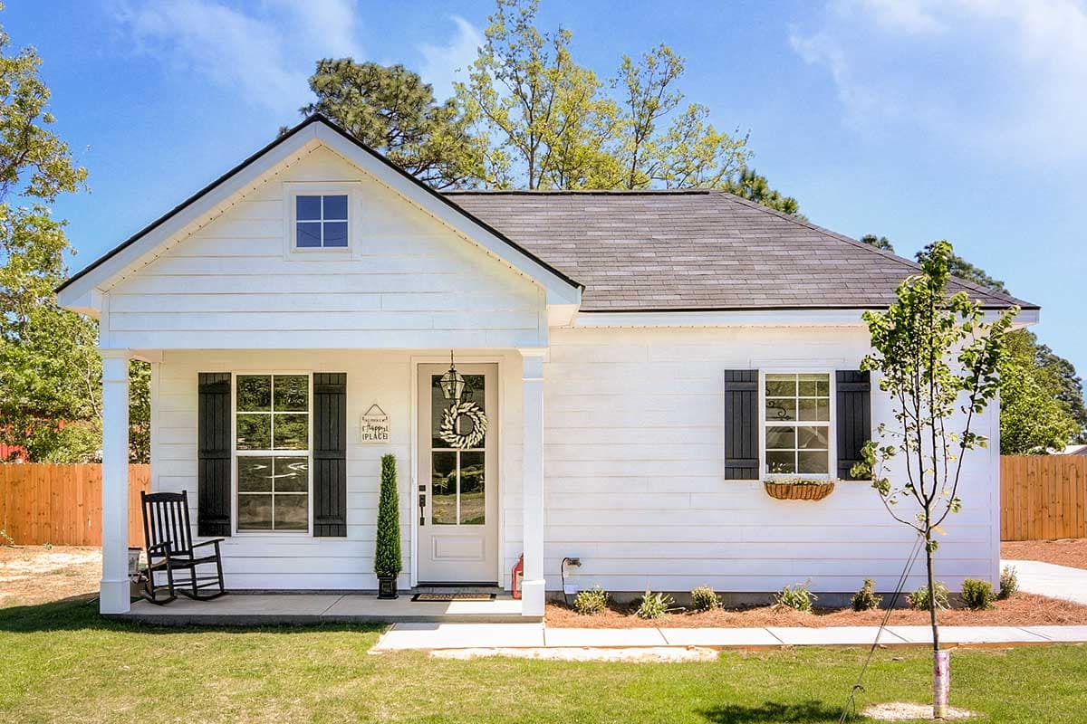 House plan exterior. Cottage style with a front porch, gable roof, and black shutters on the windows. One-story home.