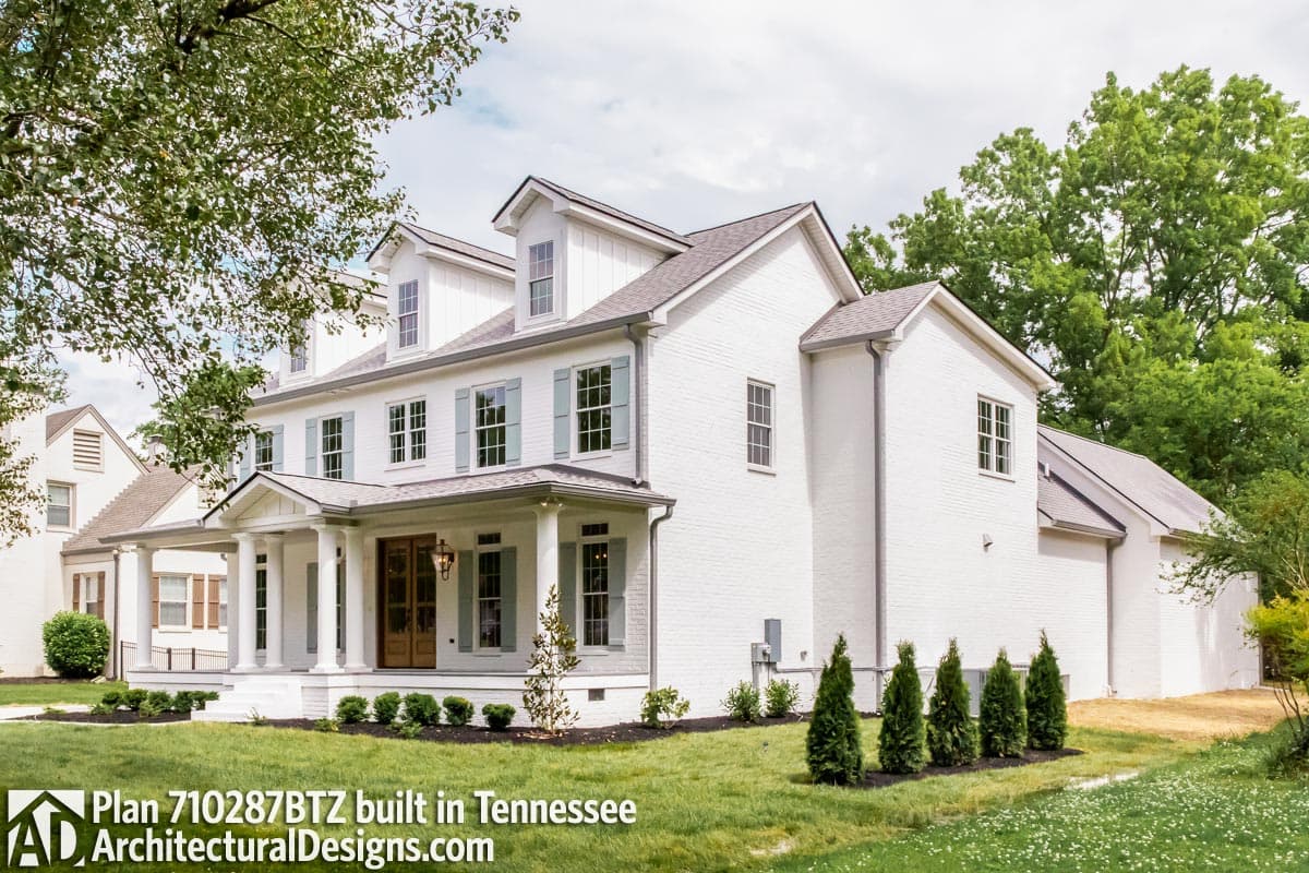 House plan exterior: Two-story white brick home with gabled dormers, prominent front porch with columns, and light blue shutters.