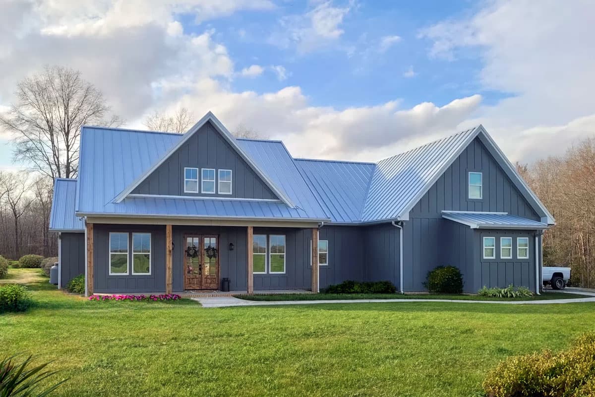 A gray farmhouse with a metal roof under a cloudy sky. The house features a porch, wooden doors, and a well-manicured lawn with a truck parked nearby.