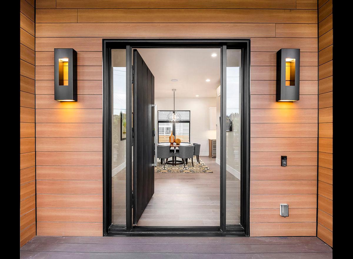 Modern house exterior with wood siding, large pivot door, and glass sidelights revealing dining area.