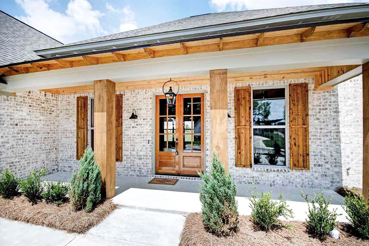 House plan exterior with white brick, wooden double doors, gabled roofline, exposed rafters, and wood-shuttered windows.
