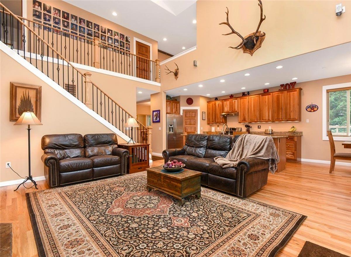 Interior view of a great room with a two-story ceiling, open staircase, leather sofas, and an adjacent kitchen with wood cabinetry.