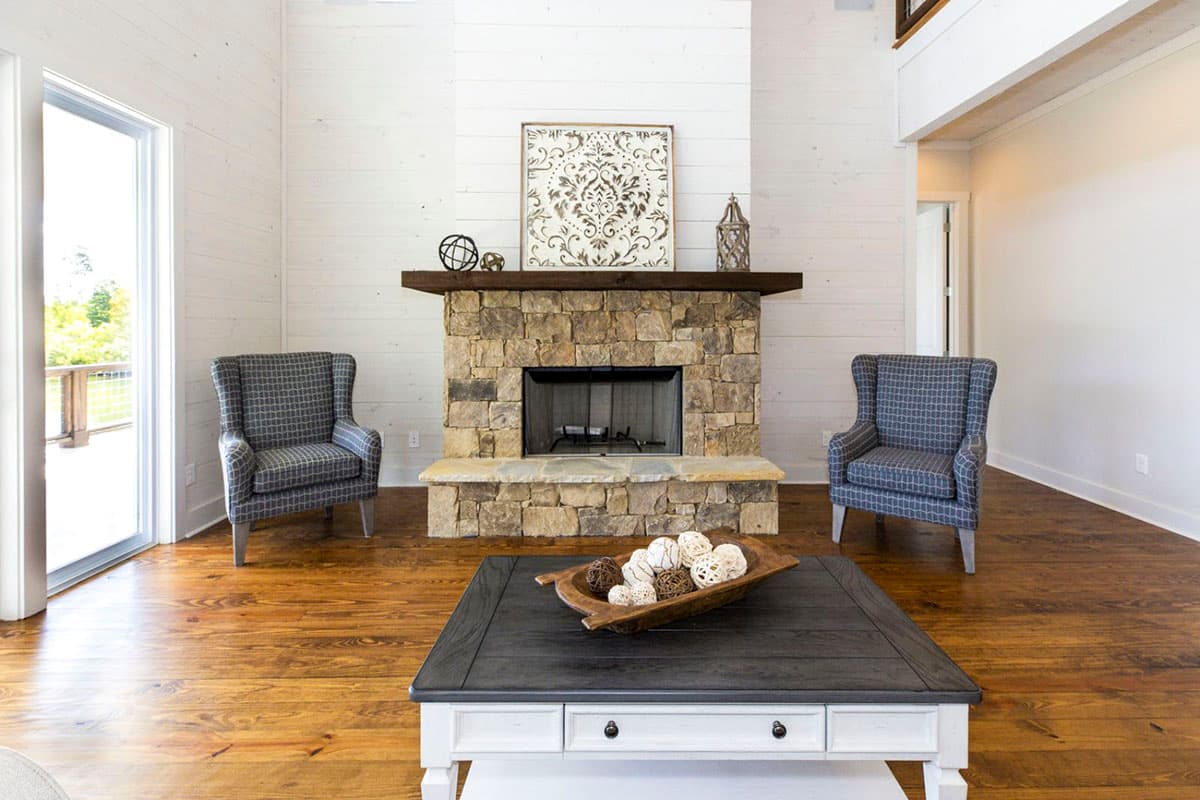 Living room with stone fireplace, vaulted shiplap ceiling, wood floors, large windows and two wingback chairs.