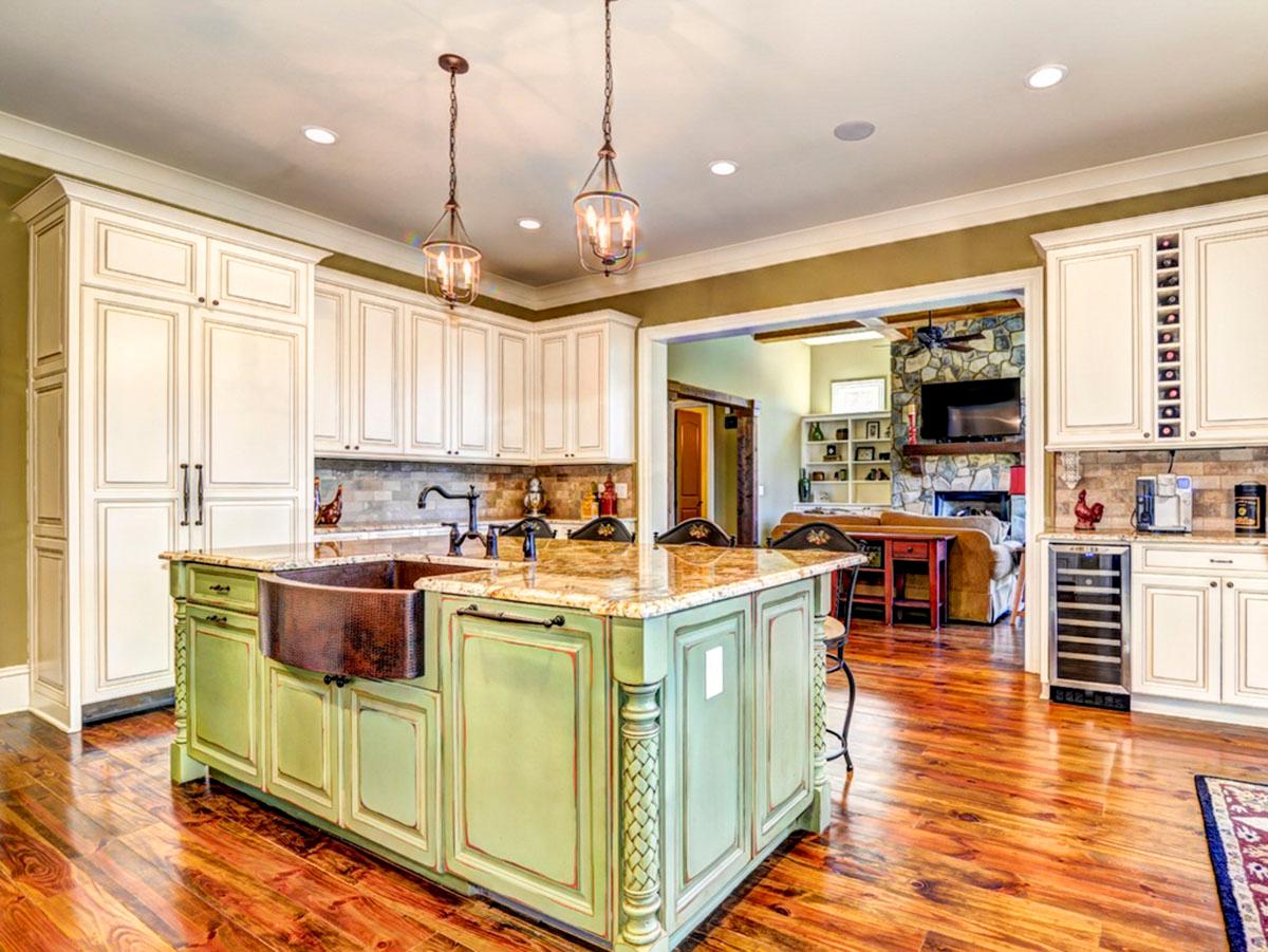 Kitchen with distressed green island, copper farmhouse sink, white cabinetry, and view into living room with stone fireplace.