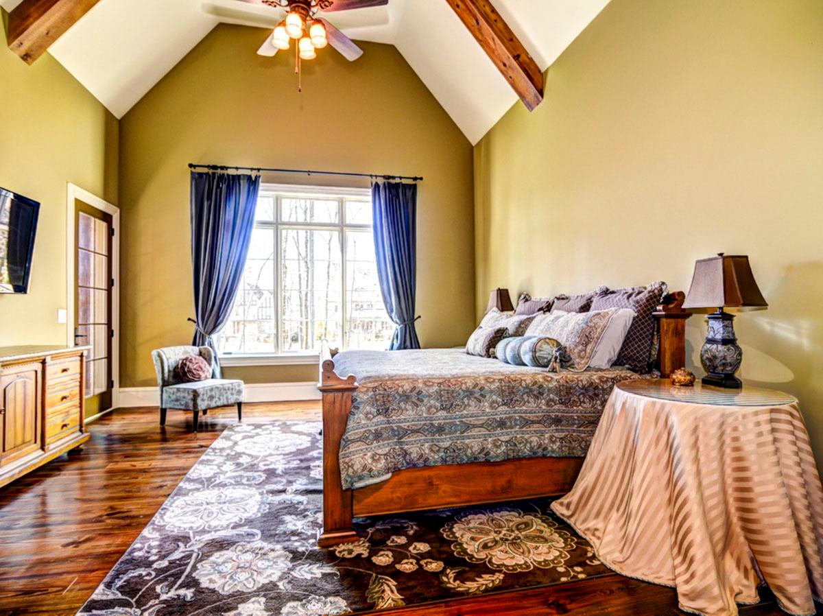 Primary bedroom interior with vaulted ceiling, exposed beams, large window, and wooden floor.