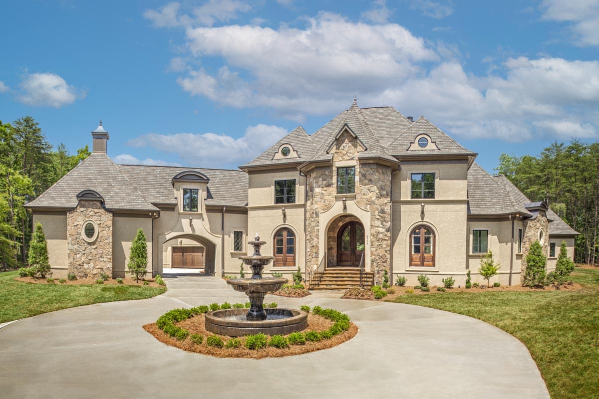 House plan exterior. Two-story home with stone accents. Features include arched entry, gabled roof, and attached garage. Driveway with fountain.