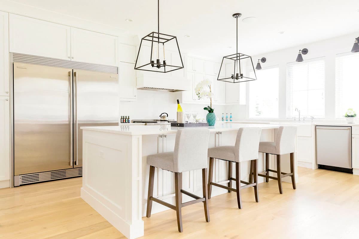 Kitchen interior with large stainless steel refrigerator, white island with seating, pendant lights, and white cabinetry.