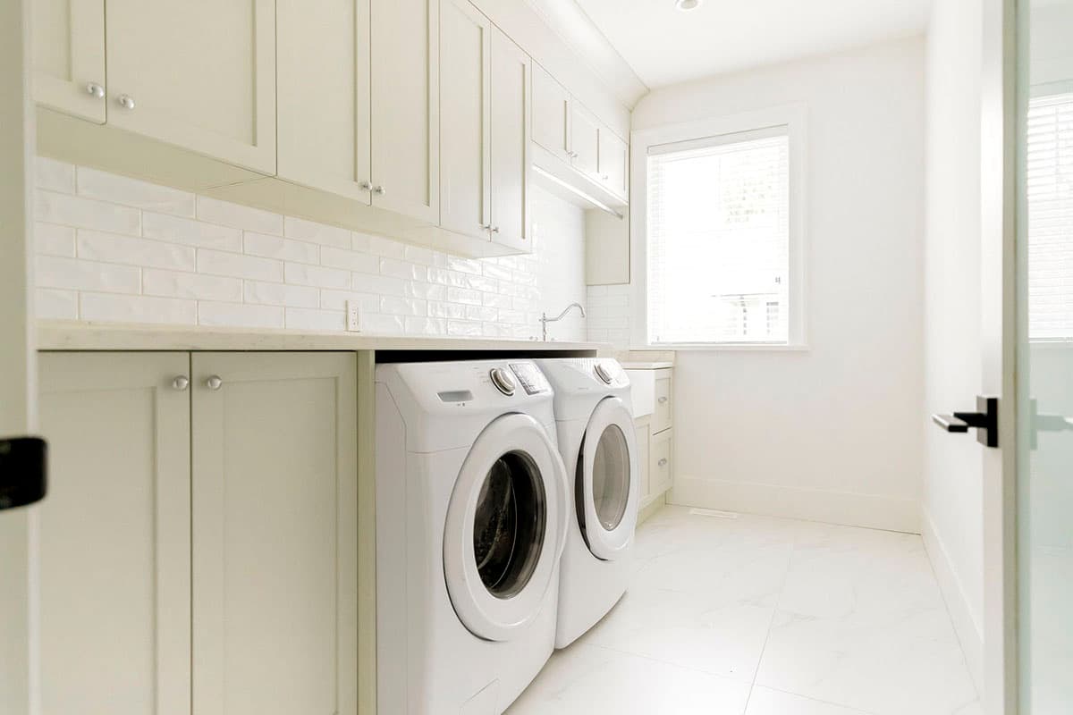 Laundry room with white subway tile backsplash, light wood cabinets, front-load washer and dryer, and white marble tile floor.