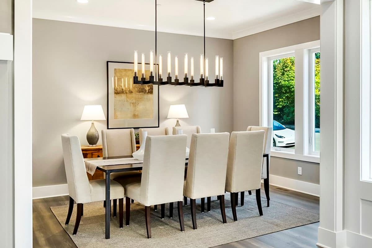Dining room with eight upholstered chairs around a dark wood table, pendant light fixture, and large window overlooking greenery.