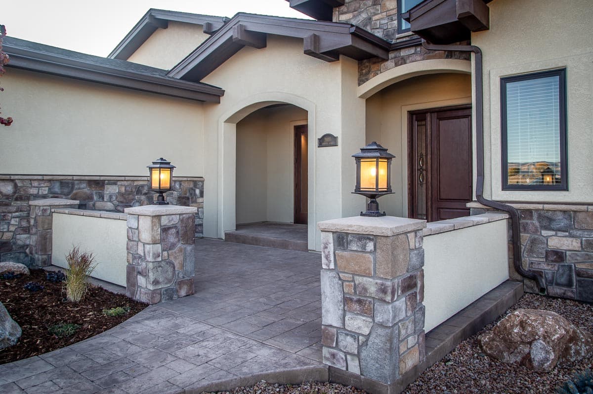House plan exterior with stone accents, arched entry, and dual carriage lamps. Gable roofline with exposed rafter tails.