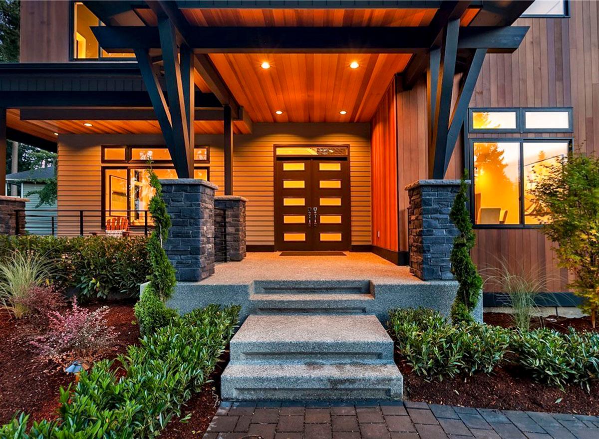 Modern house exterior featuring a covered entry with double doors, stone pillars, wood siding, and large windows.