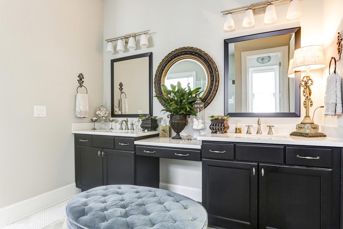 Bathroom with dual vanities, dark cabinets, white countertops, two mirrors, and a decorative ottoman.