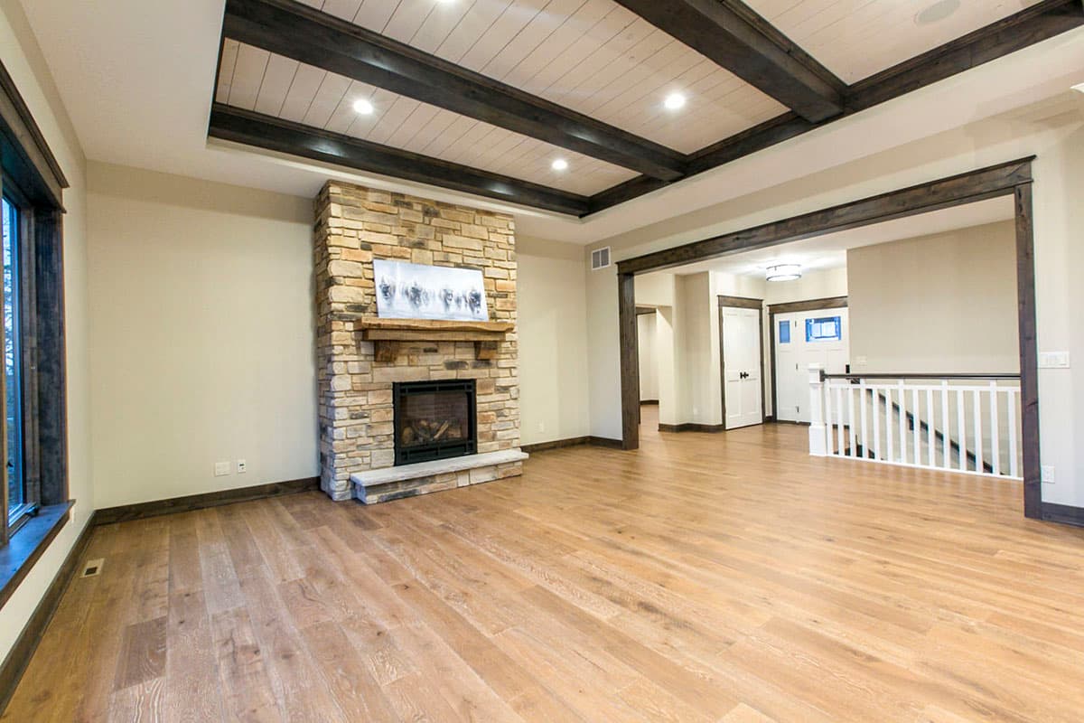 Living room interior with stone fireplace, coffered wood beam ceiling, wood floors, and view to entryway with stairs.