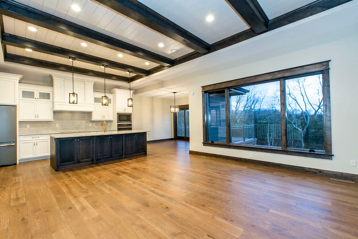 Open concept living area with kitchen island, dark wood beams on ceiling, and large windows overlooking wooded area.