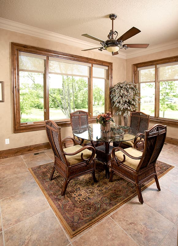 Interior view of a dining nook with a glass-top table, four wicker chairs, a ceiling fan, and large wood-trimmed windows.