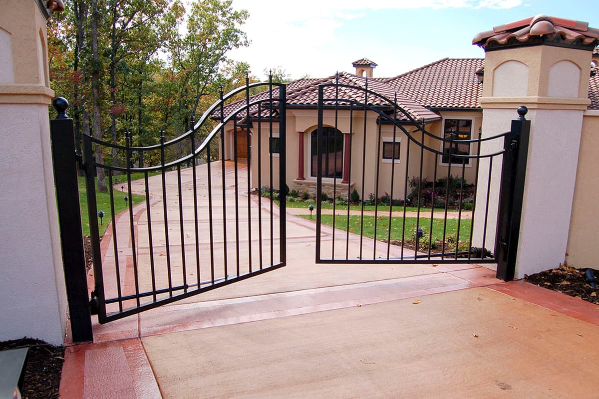 Wrought iron driveway gate opens to a light-colored stucco house with a tile roof and arched windows. House plan exterior.
