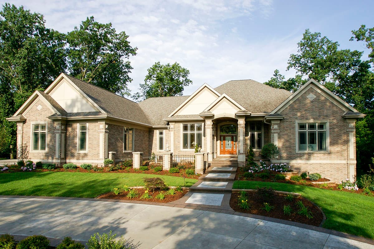 House plan exterior. Single-story brick home with gabled roof, entry portico, and multiple dormers. Landscaping includes lawn and flower beds.