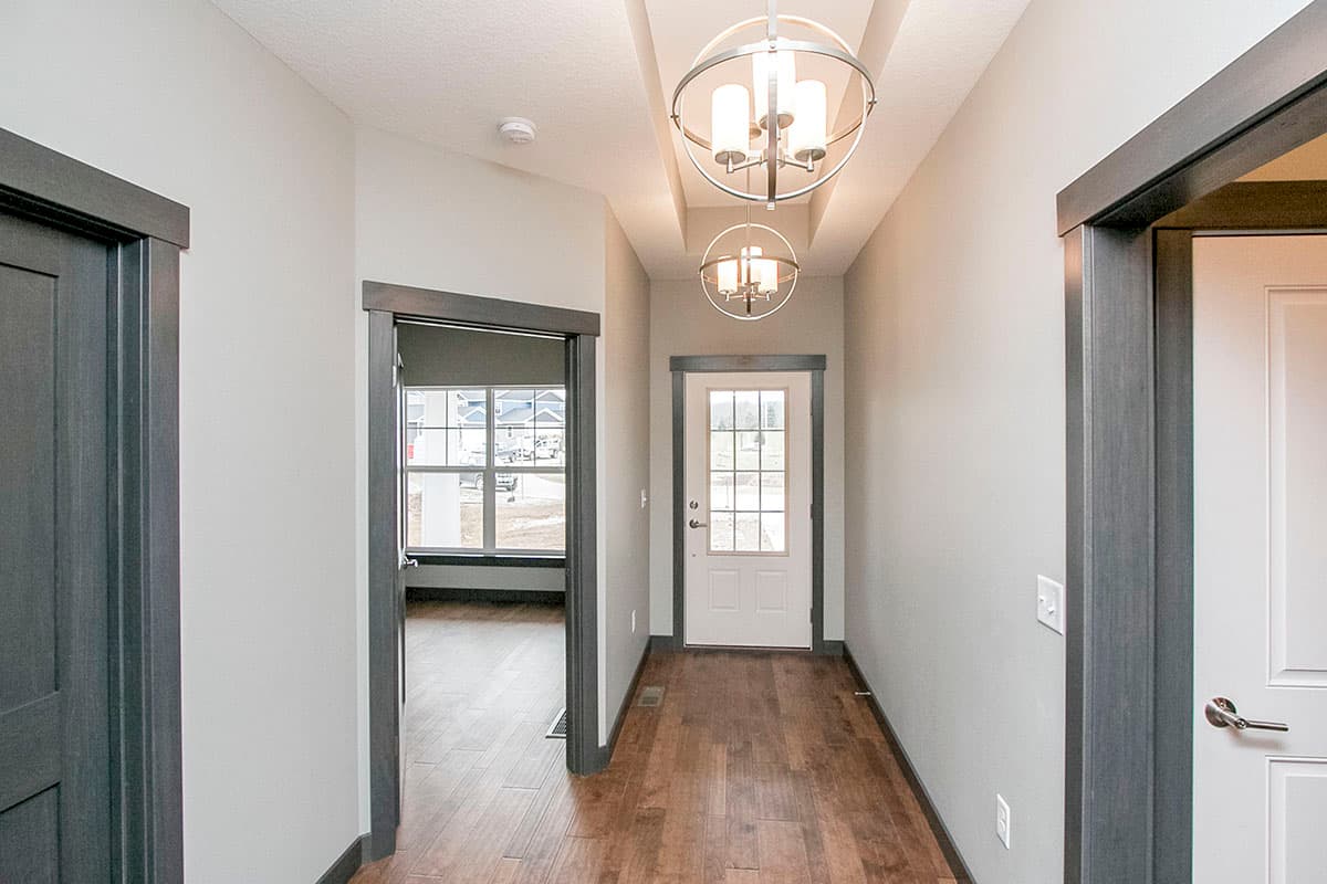 Interior view of a hallway with wood floors, leading to a front door and an open room. Features pendant lights.