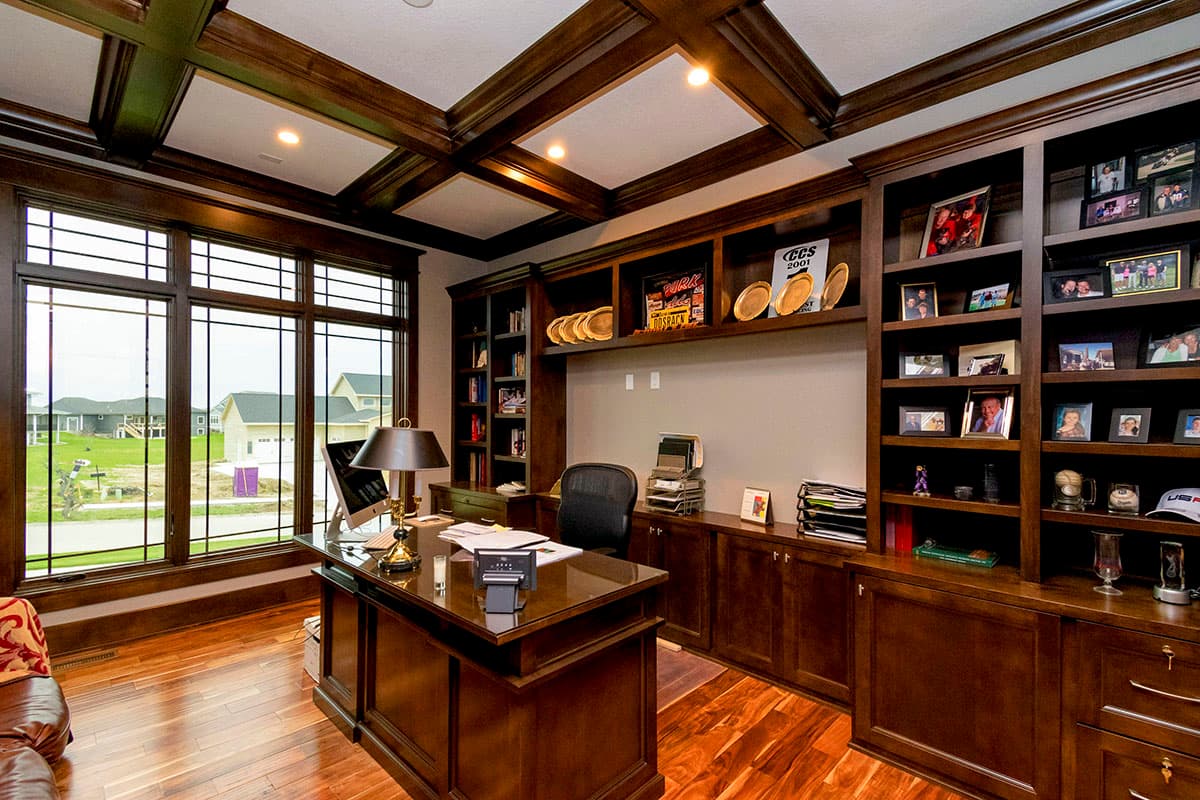 Home office with coffered ceiling, built-in dark wood bookshelves, and large grid windows overlooking a yard.