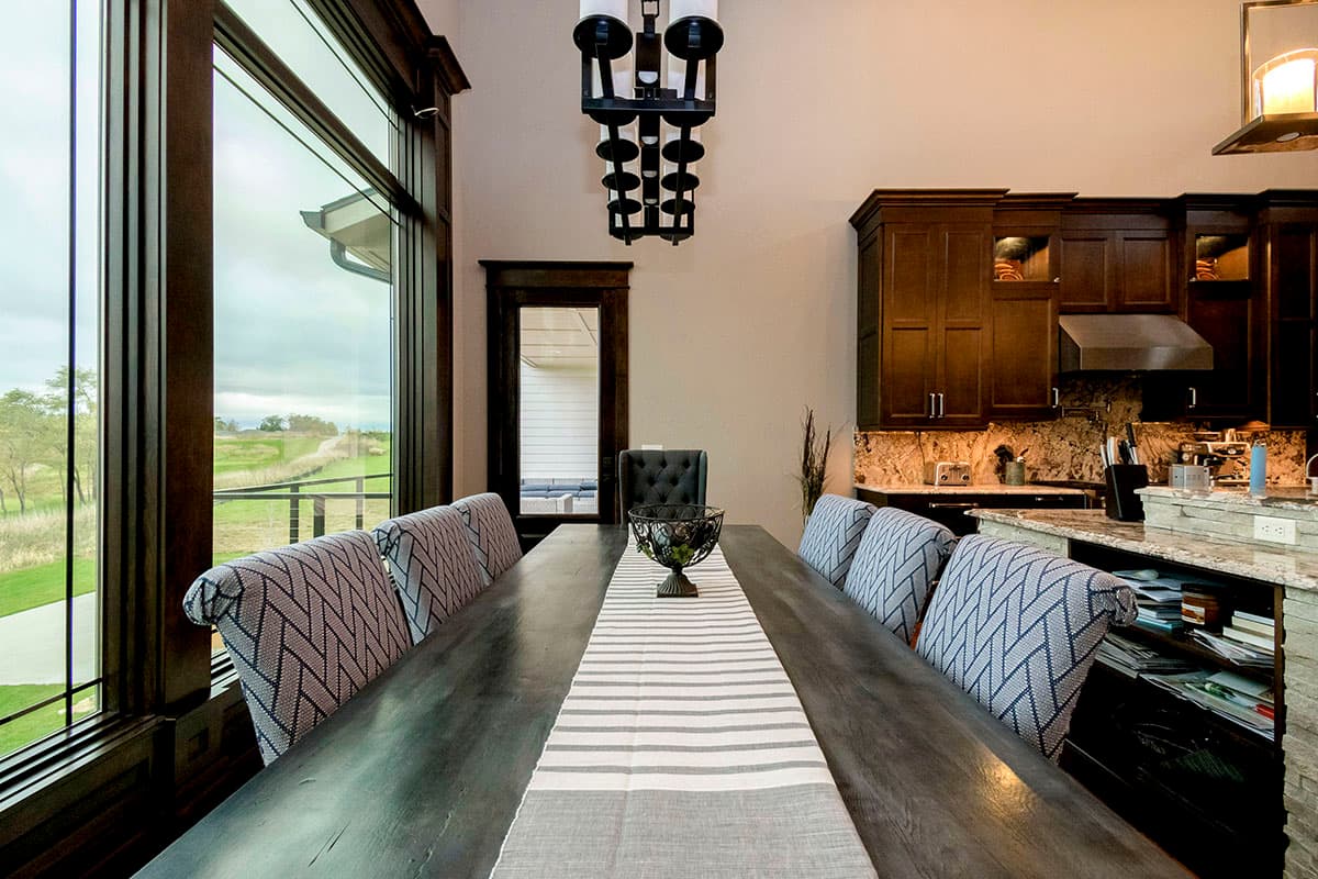 Dining room with large windows, dark wood cabinets, granite countertops, and a long wooden table with upholstered chairs.
