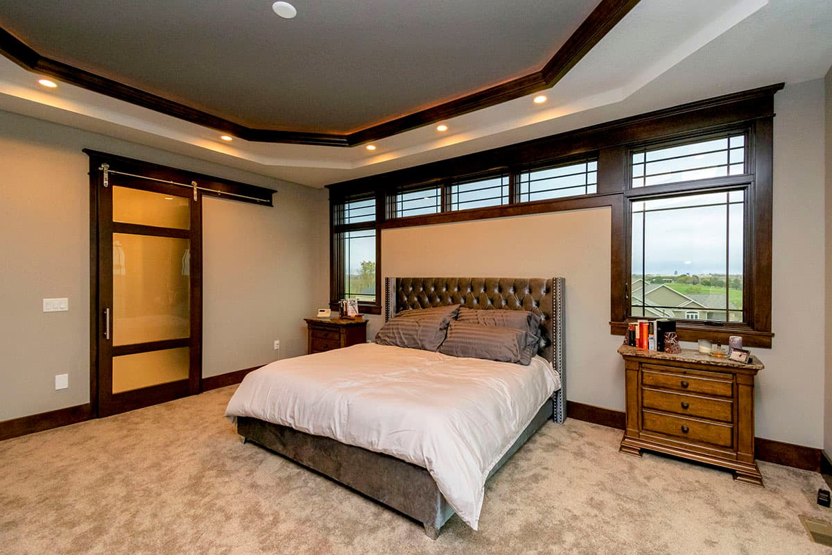 Bedroom interior with tufted headboard, sliding barn door, tray ceiling with recessed lighting, and large grid windows.
