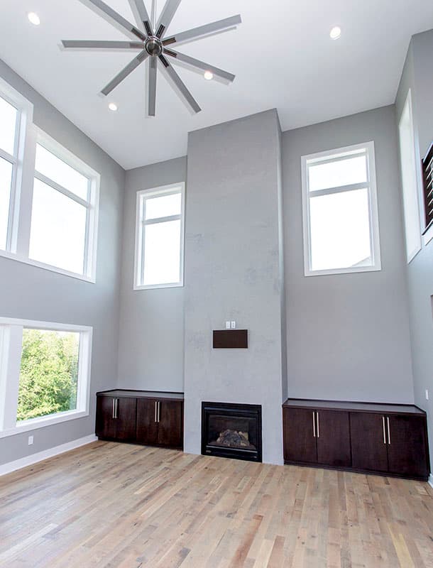 Modern living room with fireplace, built-in cabinets, and a large ceiling fan. Tall windows offer natural light.