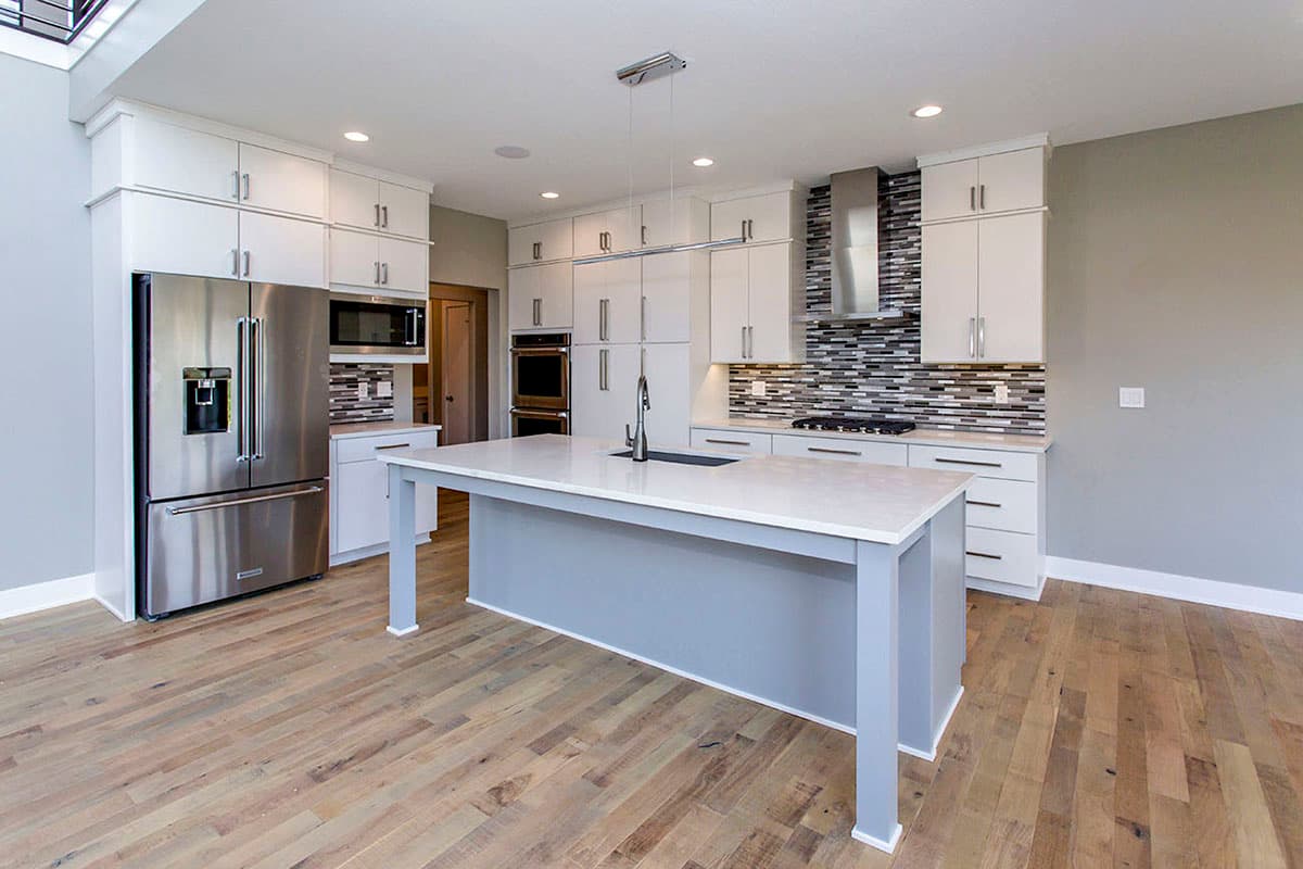 Modern kitchen interior with white cabinetry, stainless steel appliances, a large island with sink, and a mosaic tile backsplash.