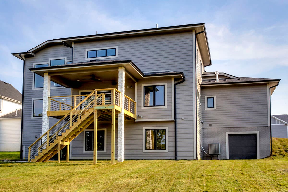 Modern house plan exterior with gray siding, a covered porch, and a wooden deck with stairs. Features a two-car garage and multiple gables.