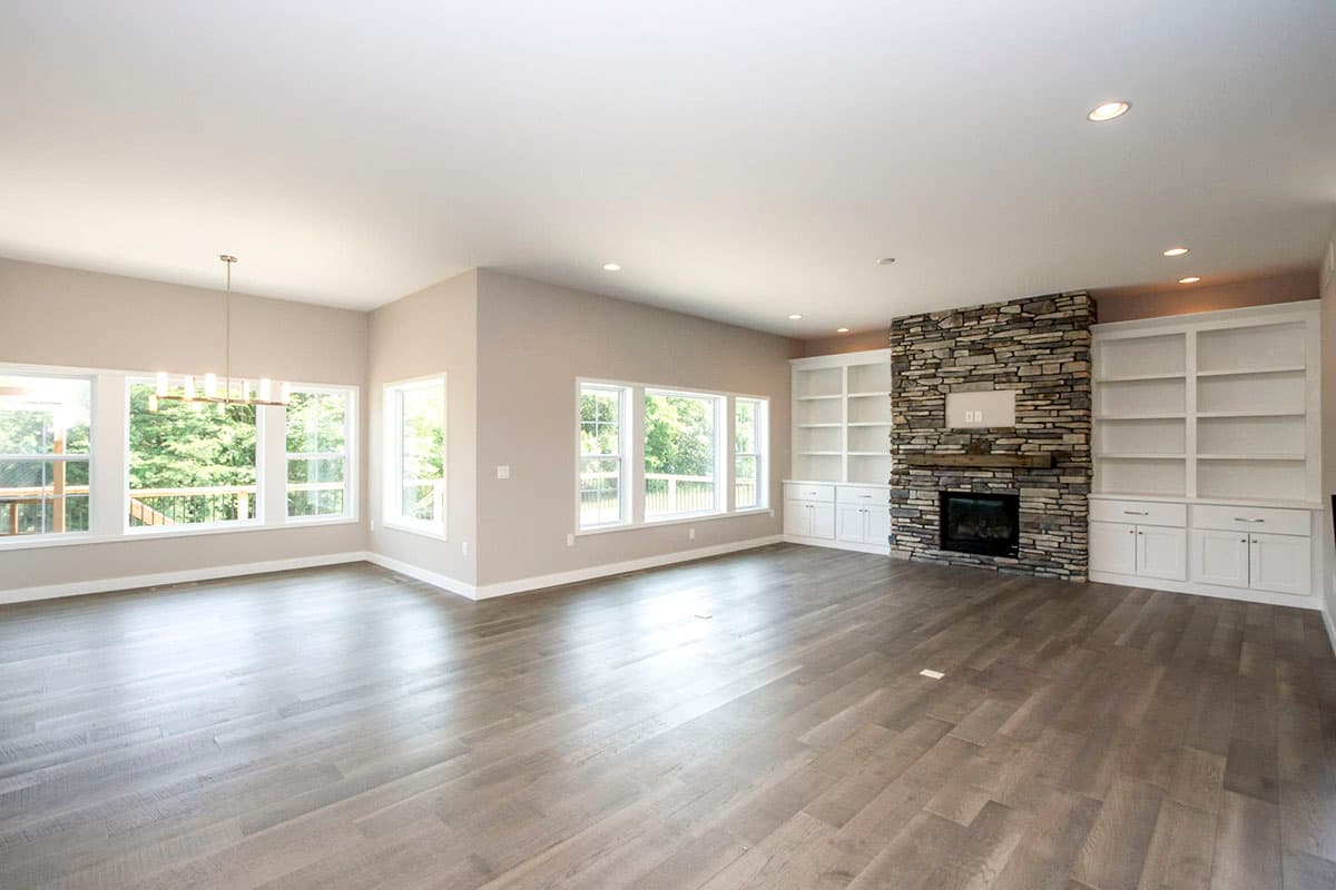 Interior view of a great room with stone fireplace, built-in bookshelves, large windows, and wood flooring.