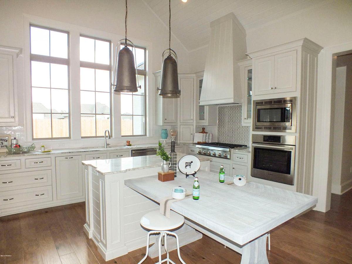 Kitchen interior with vaulted ceiling, large windows, island, stainless steel appliances, and white cabinetry.