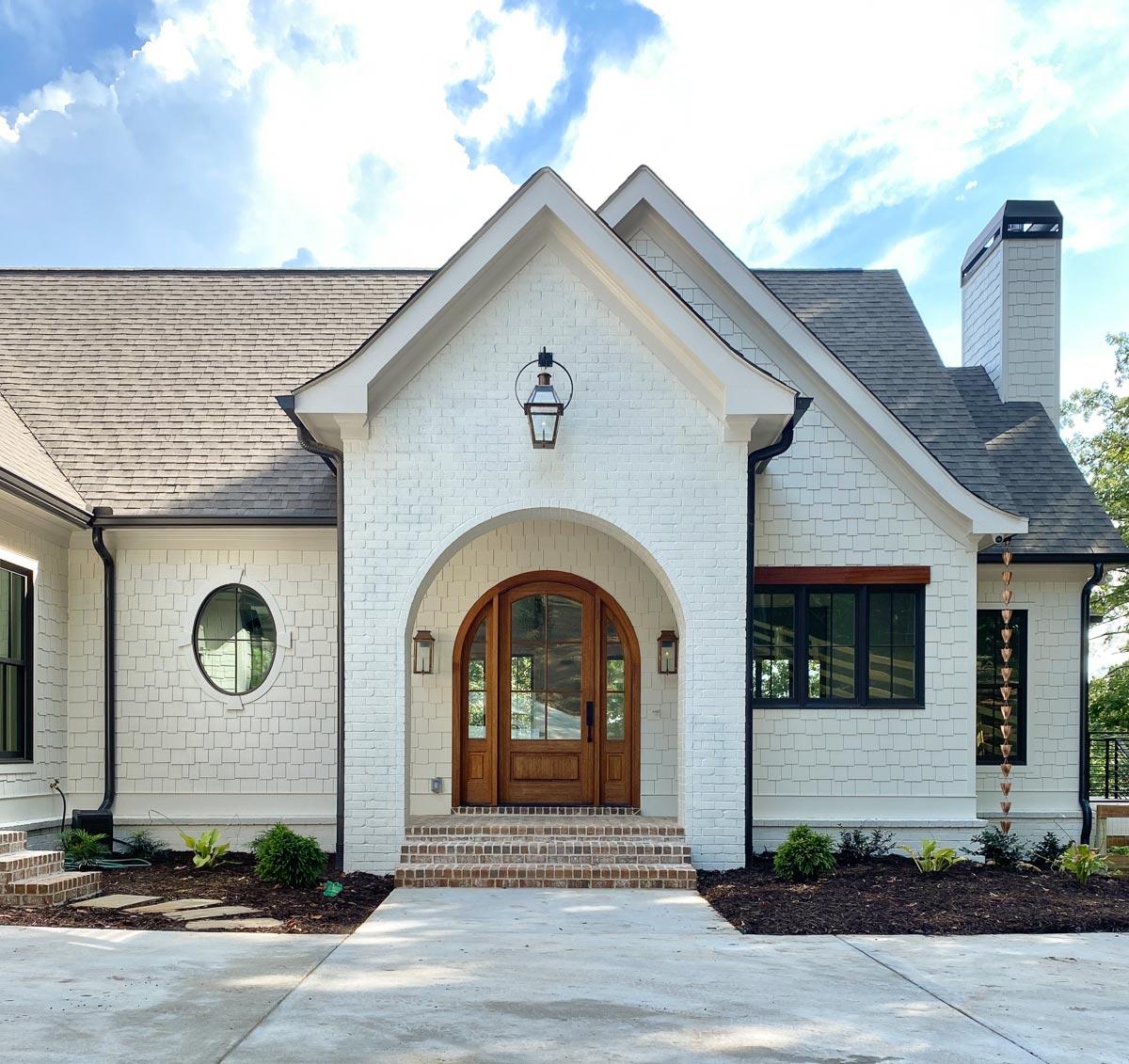 House plan exterior with white brick and shake siding, arched entry with double wooden doors, and a prominent gable.