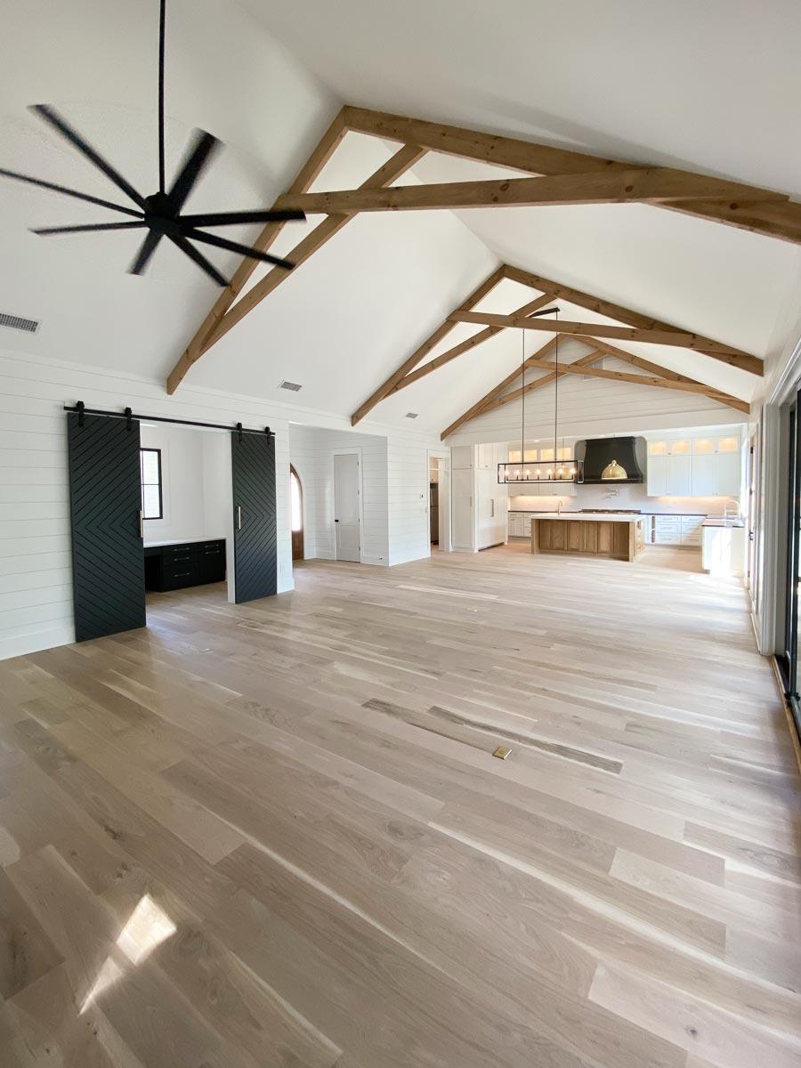 Interior view of open great room with vaulted beamed ceiling, wood floors, shiplap walls, and sliding barn doors leading to kitchen.
