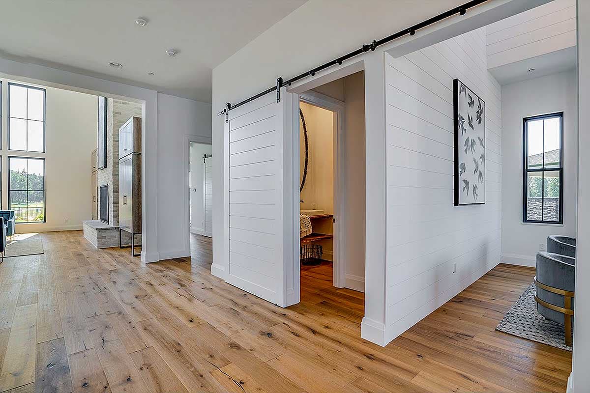 Hallway with shiplap accent wall, open doorway revealing powder room, and view of great room with fireplace and tall windows.