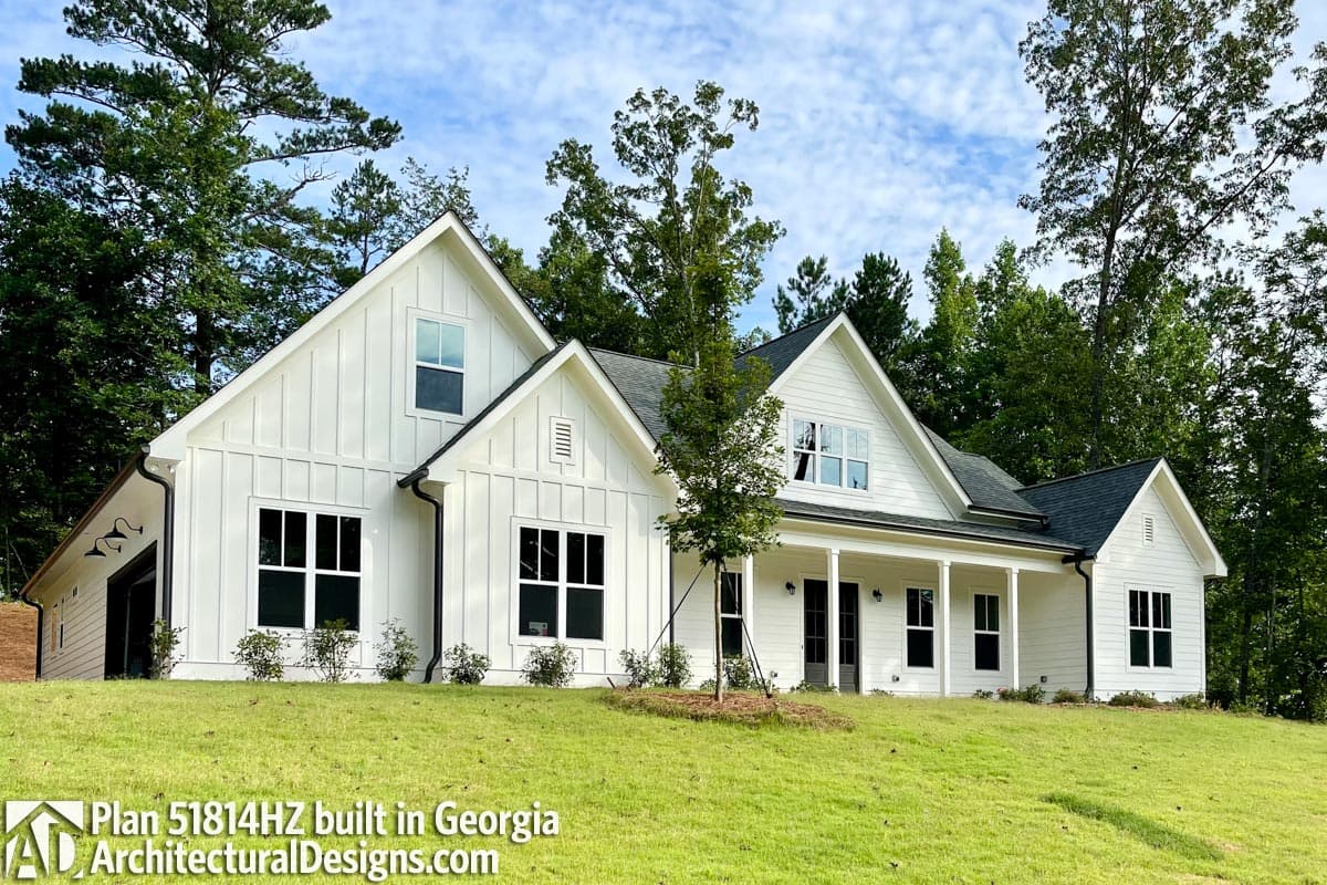 A modern, white farmhouse-style house with black accents and windows sits on a grassy hill, surrounded by trees under a blue sky.