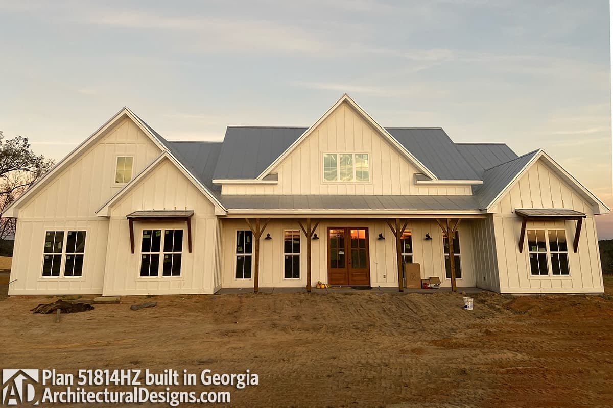A newly constructed white farmhouse with a metal roof and a large covered porch. The house is under construction, set against a soft, evening sky.