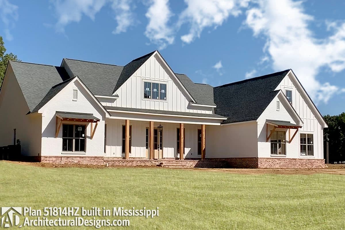 A newly constructed white farmhouse with a large front porch and brick accents. The home sits on a grassy lawn under a partly cloudy sky.