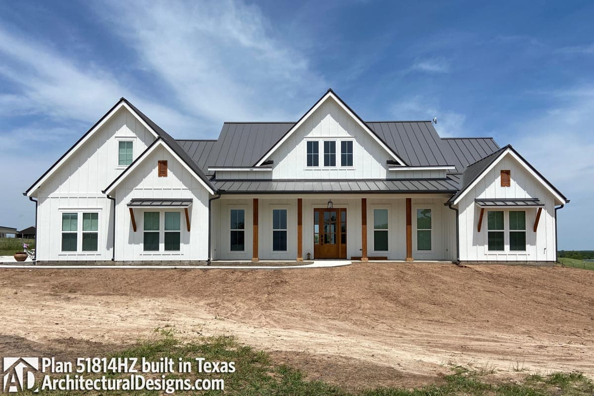 A newly constructed white farmhouse with a grey metal roof and wooden porch pillars under a bright blue sky. The house sits on a freshly graded lot.