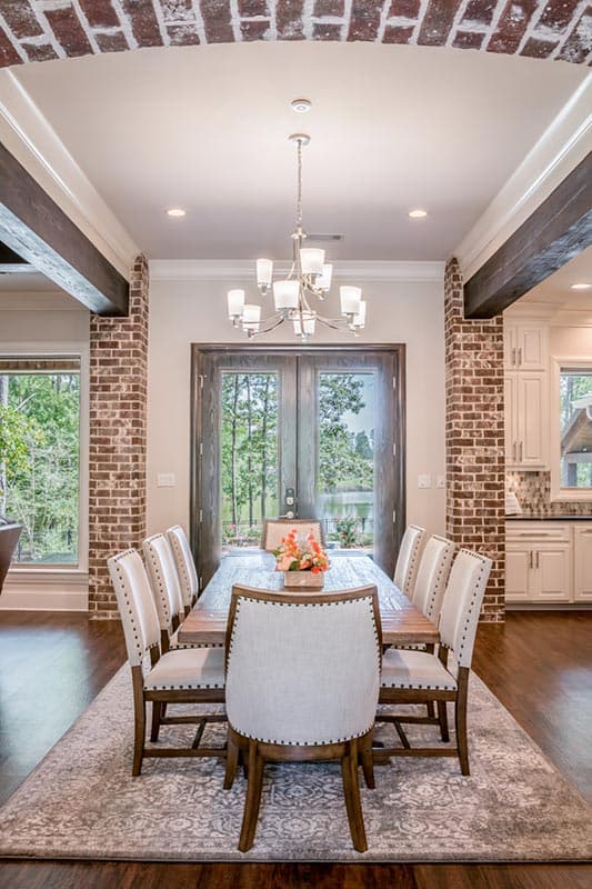 Formal dining room with long wooden table, eight upholstered chairs, and a chandelier. A brick archway and double doors lead to a lake view.