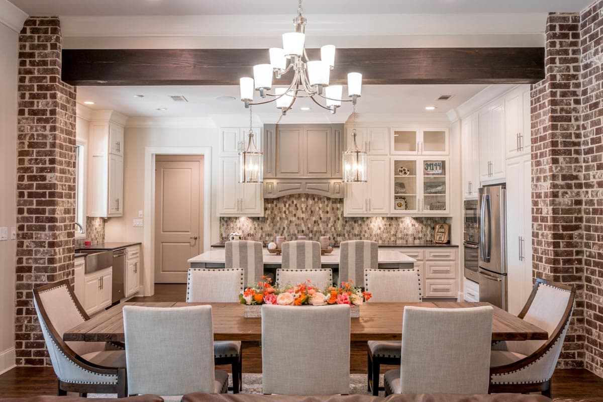Spacious dining room with a long wooden table, decorated with a floral centerpiece. The open-plan kitchen features white cabinets and brick accents.