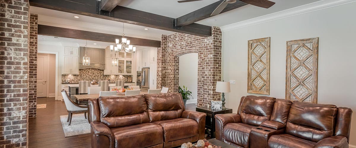 A luxury living room with leather furniture and an open kitchen. Exposed brick accents the archway to the kitchen and the structural beams.