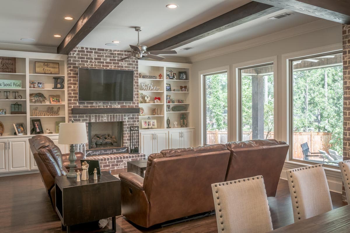 A cozy living room with a brick fireplace, built-in shelves, leather sofas, and large windows overlooking a backyard.