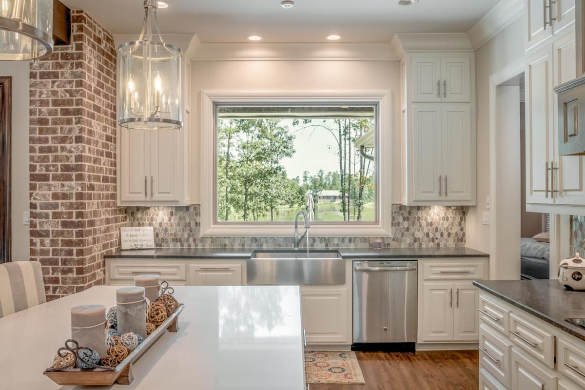 A modern kitchen features white cabinets, a stainless steel sink, and a large window overlooking a lake and trees. A brick accent wall is present.