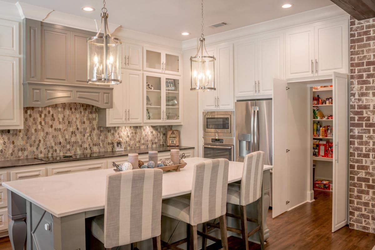 A modern kitchen with a large island, white cabinets, stainless steel appliances, and a pantry with the door open revealing contents.